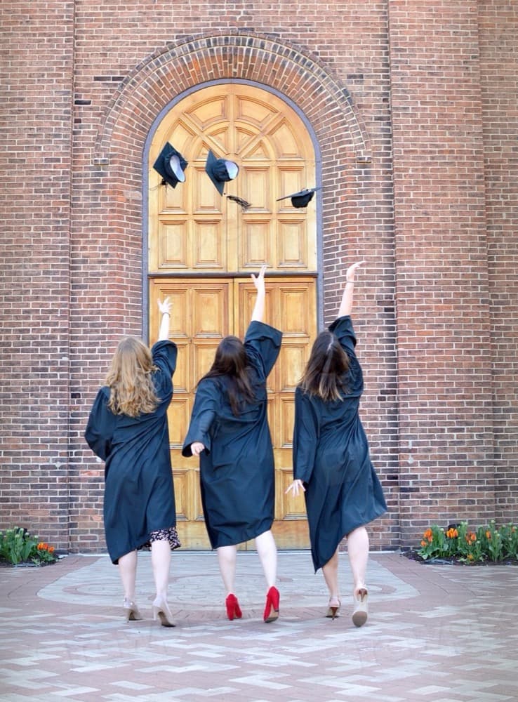 Three graduates in gowns throwing their caps in the air outside a school entrance.
